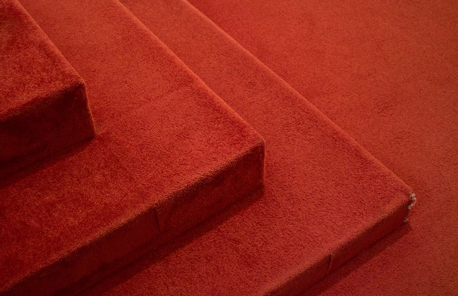 Close-up view of a red carpeted staircase in a residential setting, showing three visible steps with a plush texture and consistent color. The carpet appears clean and well-maintained, with minimal si